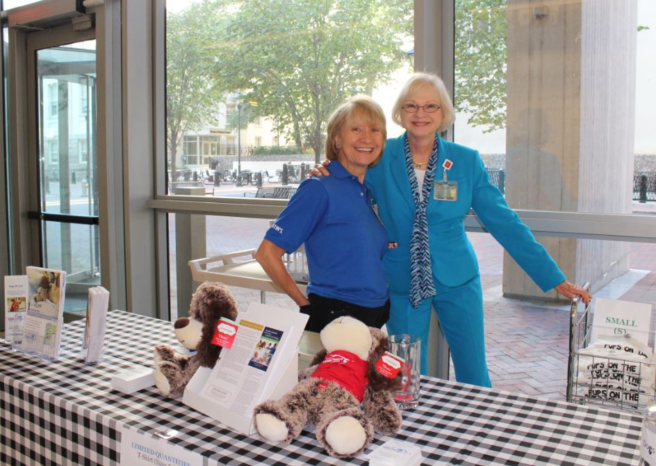 Dr. Sandra Barker and a Dogs On Call volunteer pose in front of a table at pups on the plaza 2017