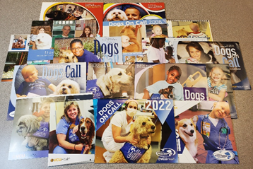 A pile of roughly 20 calendars is spread out on a table.  The covers of the calendars are each from a different year, and the photos on the covers vary from nurses to patients to children, all loving on DOC dogs.