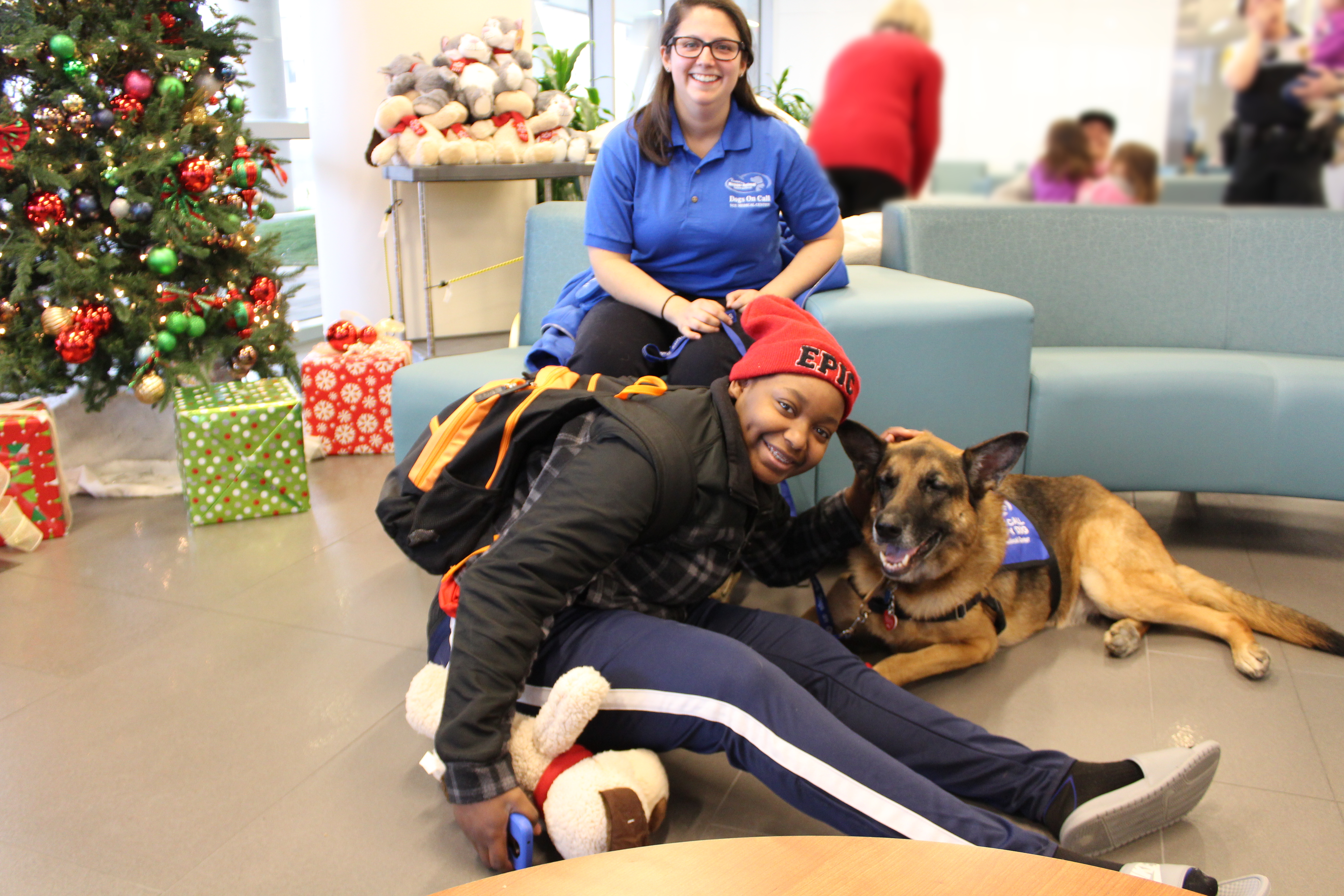 Therapy dog Dahlia, a german shepherd, lays on the floor beside a friend who is also holding a PetSmart Charities stuffed dog. Dahlia's handler sits behind them smiling.