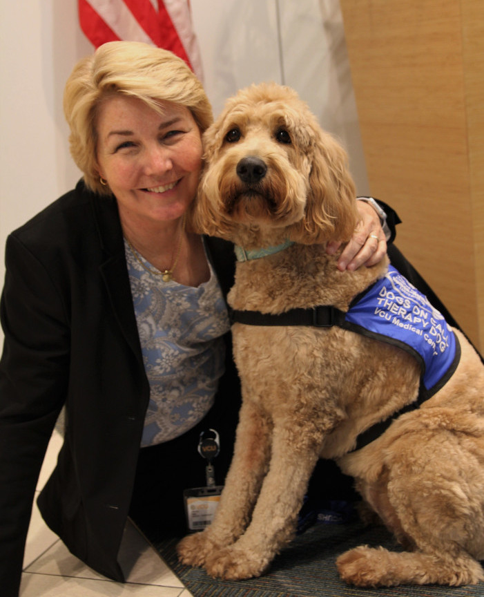 Dr. Gee sits on the floor next to Elsa, a cream-colored golden doodle wearing a blue Dogs on Call therapy dog vest