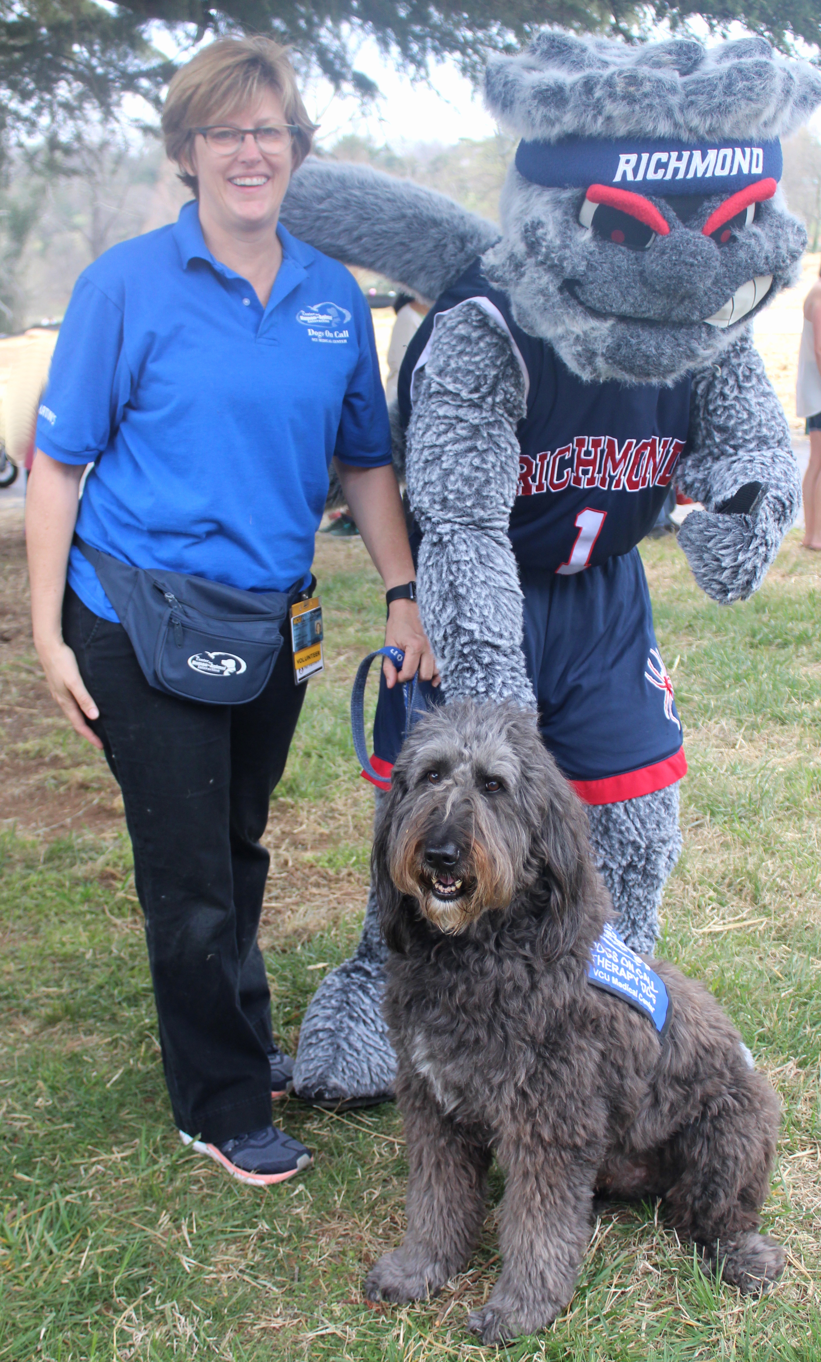 Dogs On Call therapy dog Clarke, a dark gray goldendoodle, stands in front of his handler and the University of Richmond's mascot, Webster.