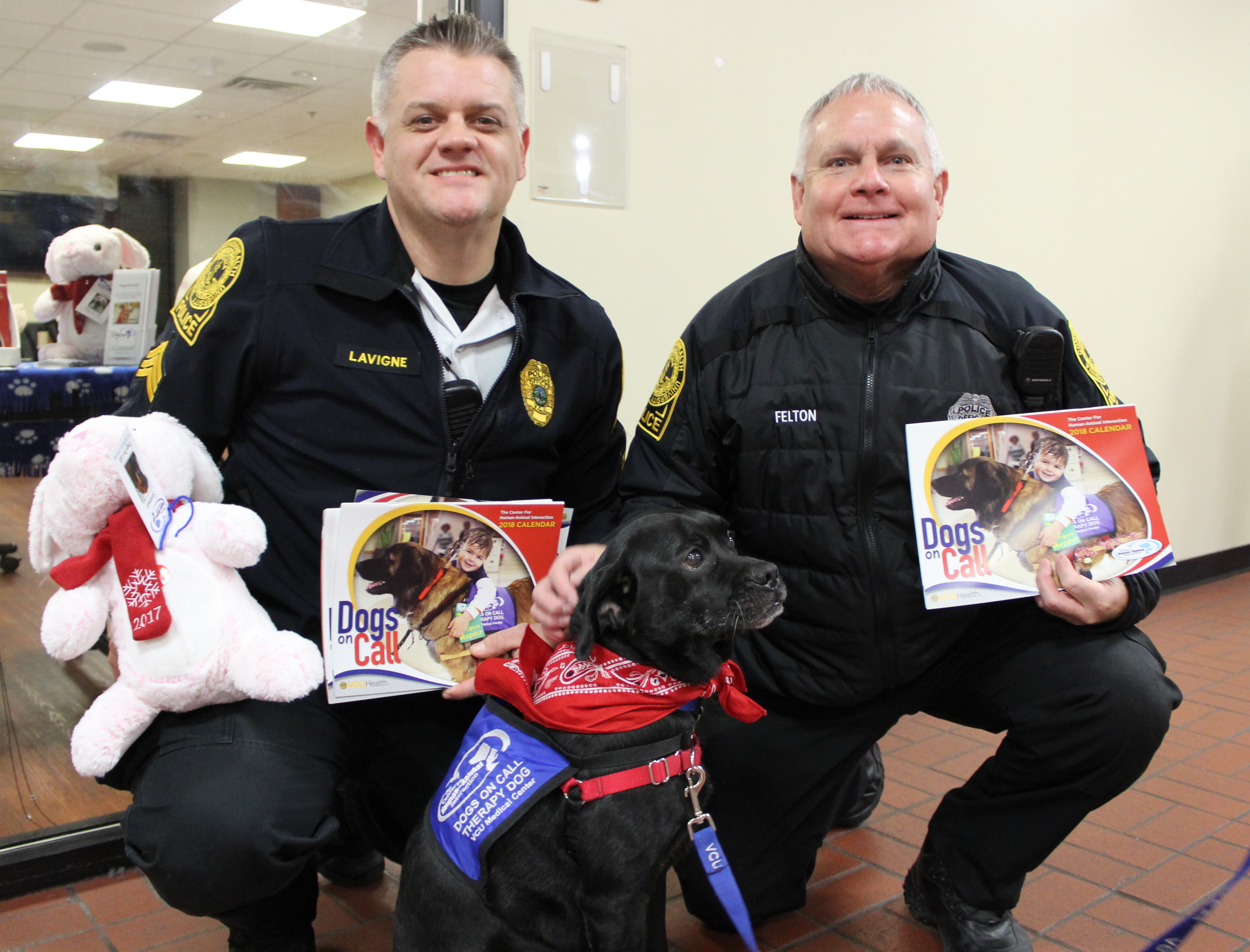 Two V C U police officers kneel beside Otis, a black lab and pug mix, while they hold the 2018 Dogs On Call Calendar and a pink stuffed animal donated by PetSmart Charities