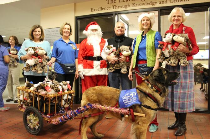 Dogs On Call therapy dog Kepler, a leonberger, pulls a cart behind her with therapy dog Phoebe, a maltipoo, sitting among stuffed animals. Dr. Sandra Barker is joined by four volunteers and Santa Claus. They are also holding stuffed animals.