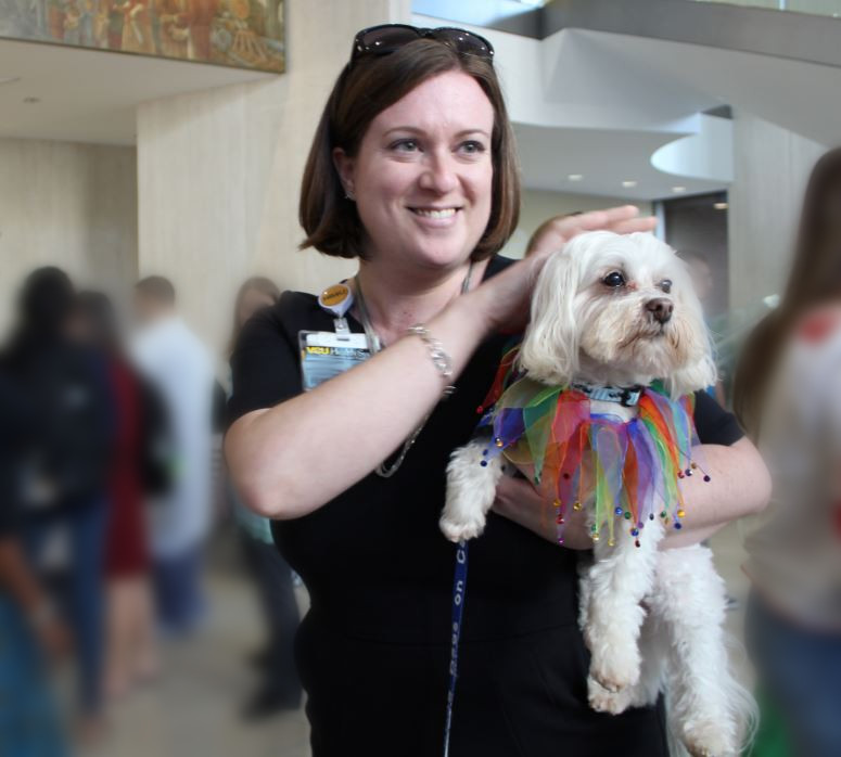 Dogs On Call therapy dog Stewie, a white maltese, is wearing a multi-colored ribbon color as he is held by a fan who is smiling as she holds him.
