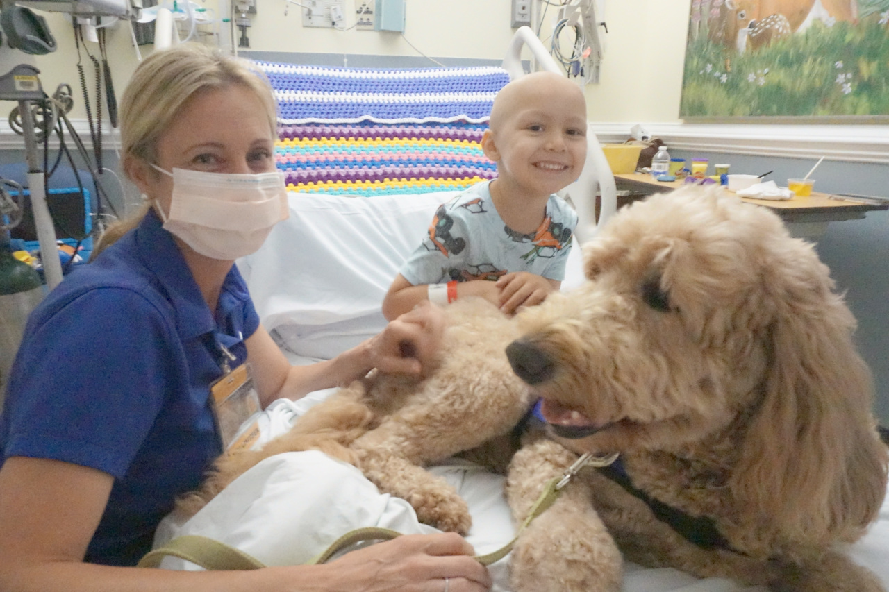 A Golden Doodle sits on the bed of a pediatric patient.  The pediatric patient is resting his arms on the dog, and is looking and smiling at the camera.