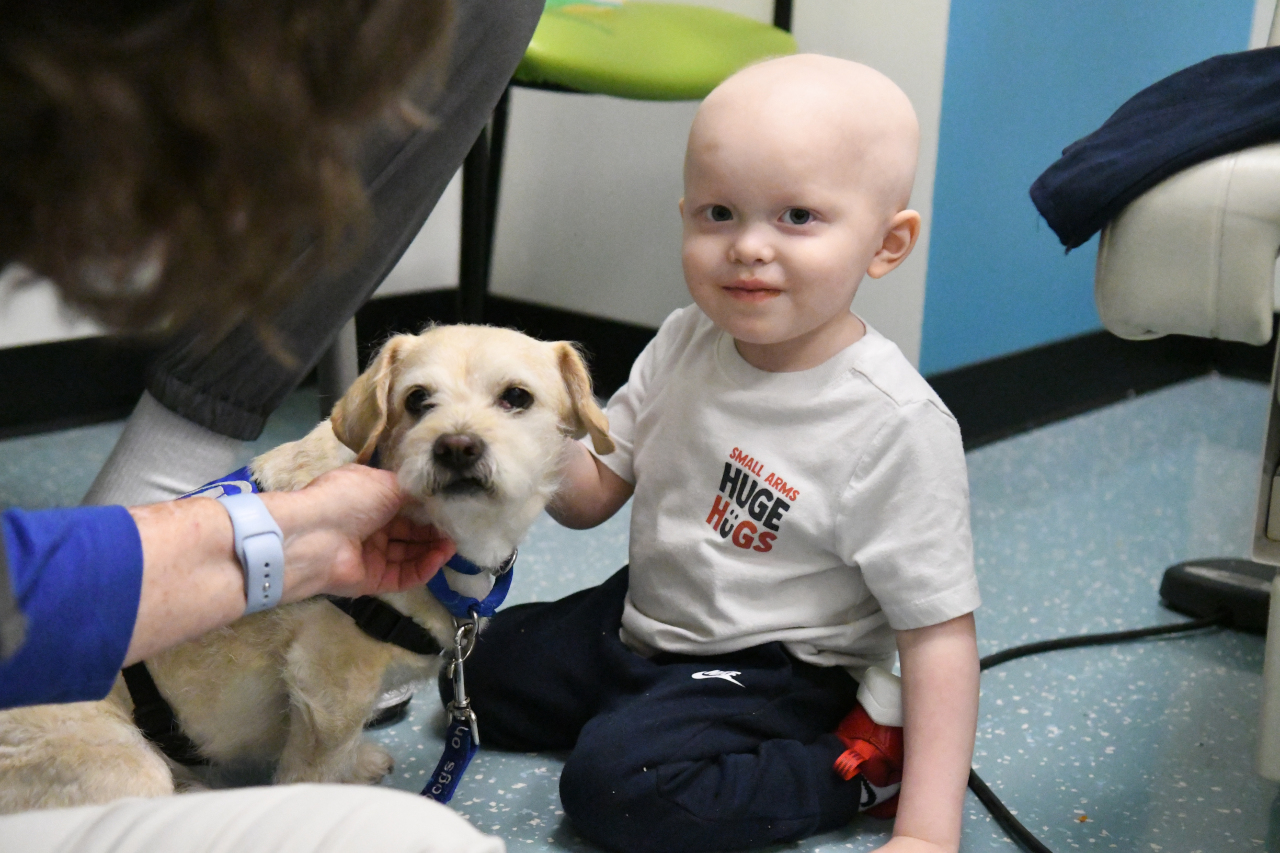 A young pediatric patient smiles at the camera as an even tinier Terrier Mixed breed dog sits next o him.