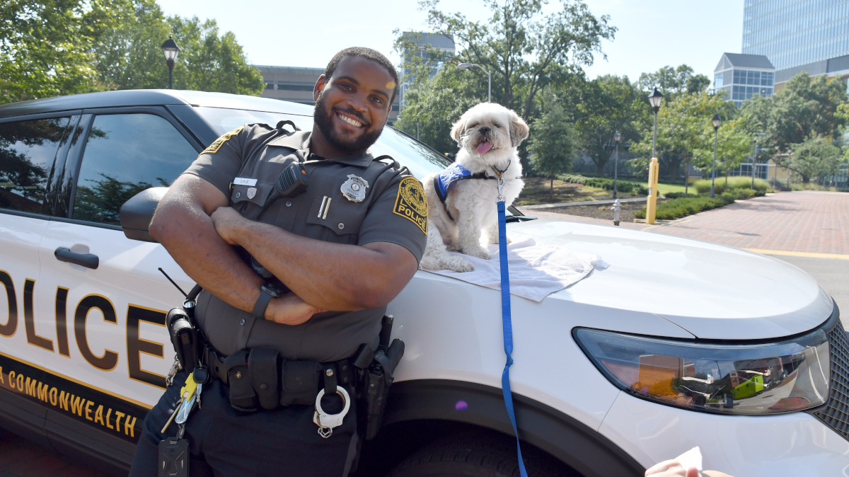 A police officer stands with his arms crossed and smiling as he leans up against the hood of a police cruiser.  A small Shih Tzu smiles at the camera while sitting on a towel on the hood of the vehicle.  VCU campus can be seen in the background.