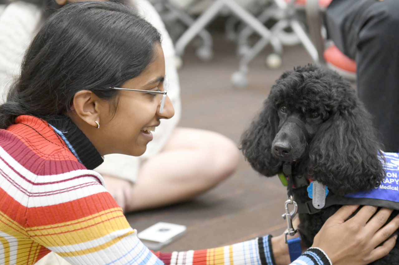 A VCU student sits on the floor while petting a black Miniature Poodle.  The student is smiling and looking at the dog, while the dog is looking at the camera.