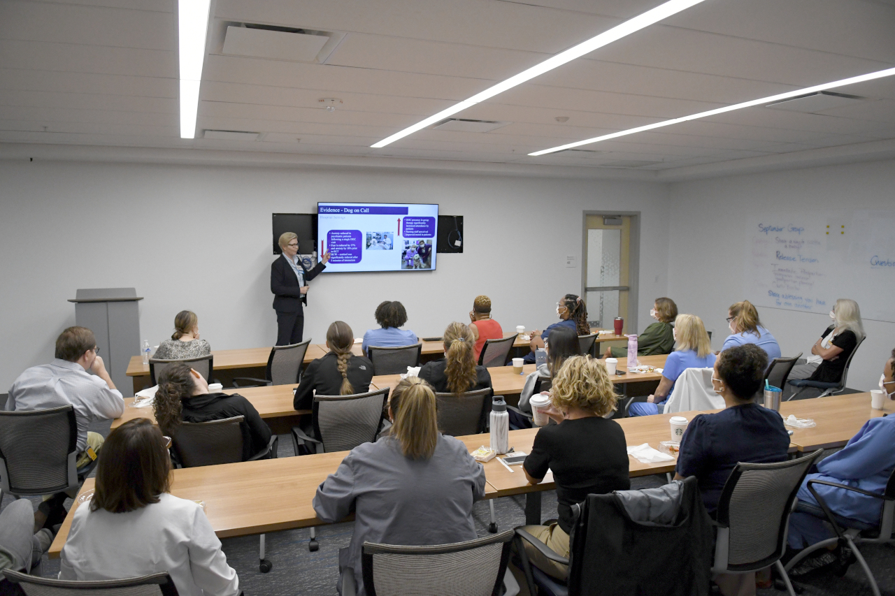 Dr. Gee stands at the front of a classroom filled with medical professional.  She gestures to a presentation on the evidence base supporting the efficacy of therapy dog visits.
