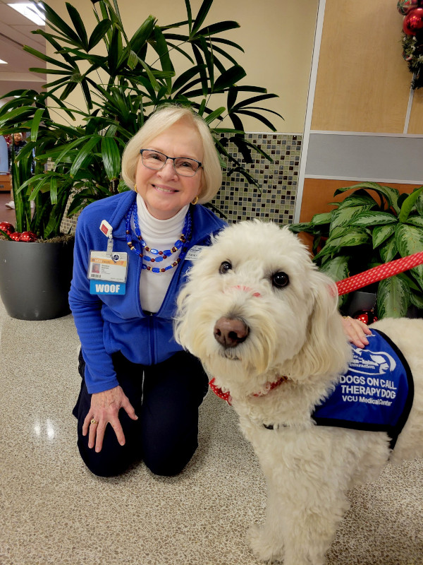 Dr. Sandra Barker kneels next to a while doodle wearing a blue Dogs on Call therapy dog vest