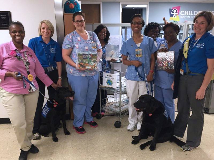 Team members from the Childrens Hospital at VCU pose with Earth Day Books and two Dogs On Call therapy dogs who are black labs and their handlers.  