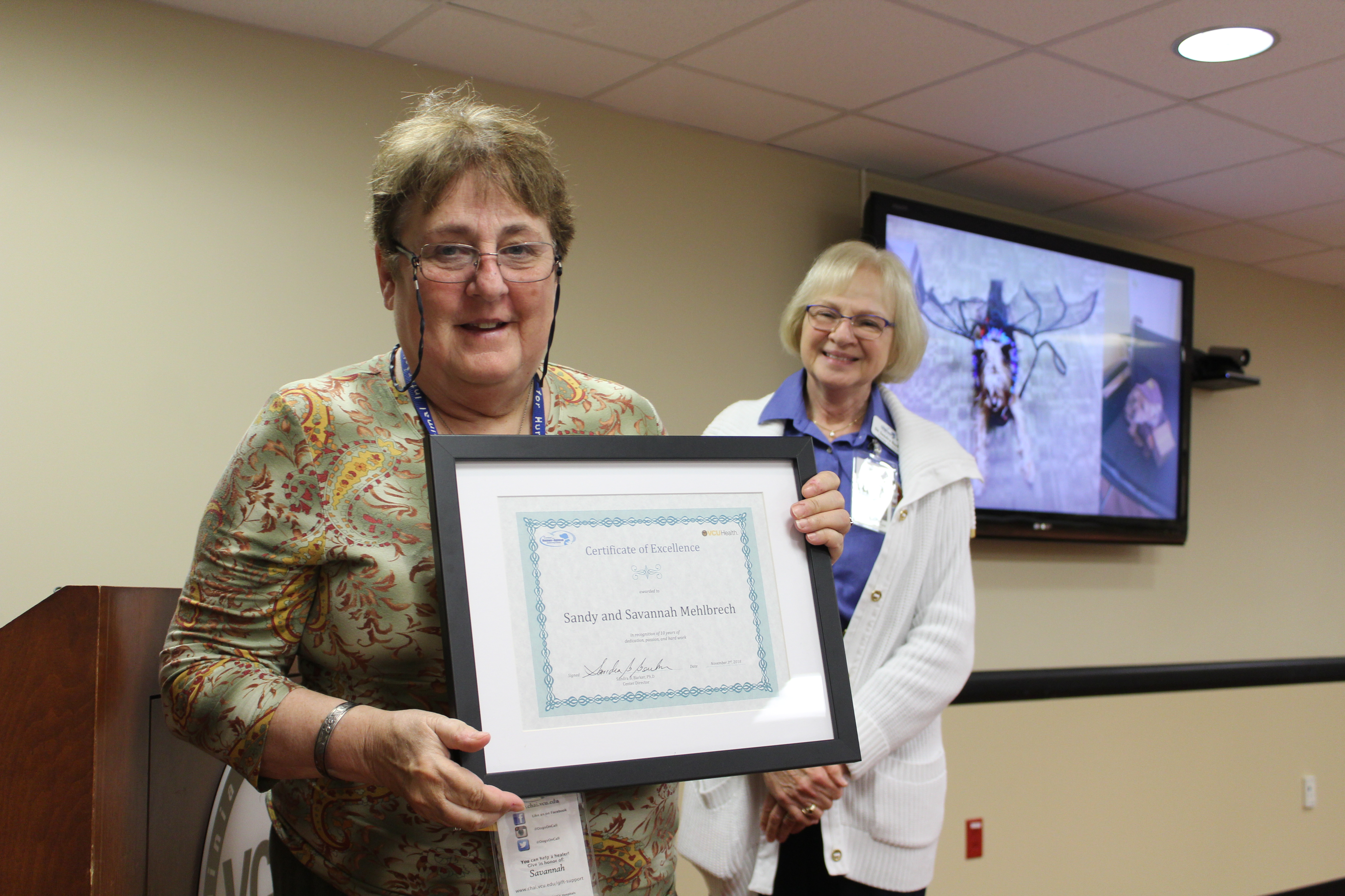 A Dogs On Call therapy dog handler receives a framed certificate recognizing 10 years of volunteering. Dr. Barker stands behind the smiling volunteer.