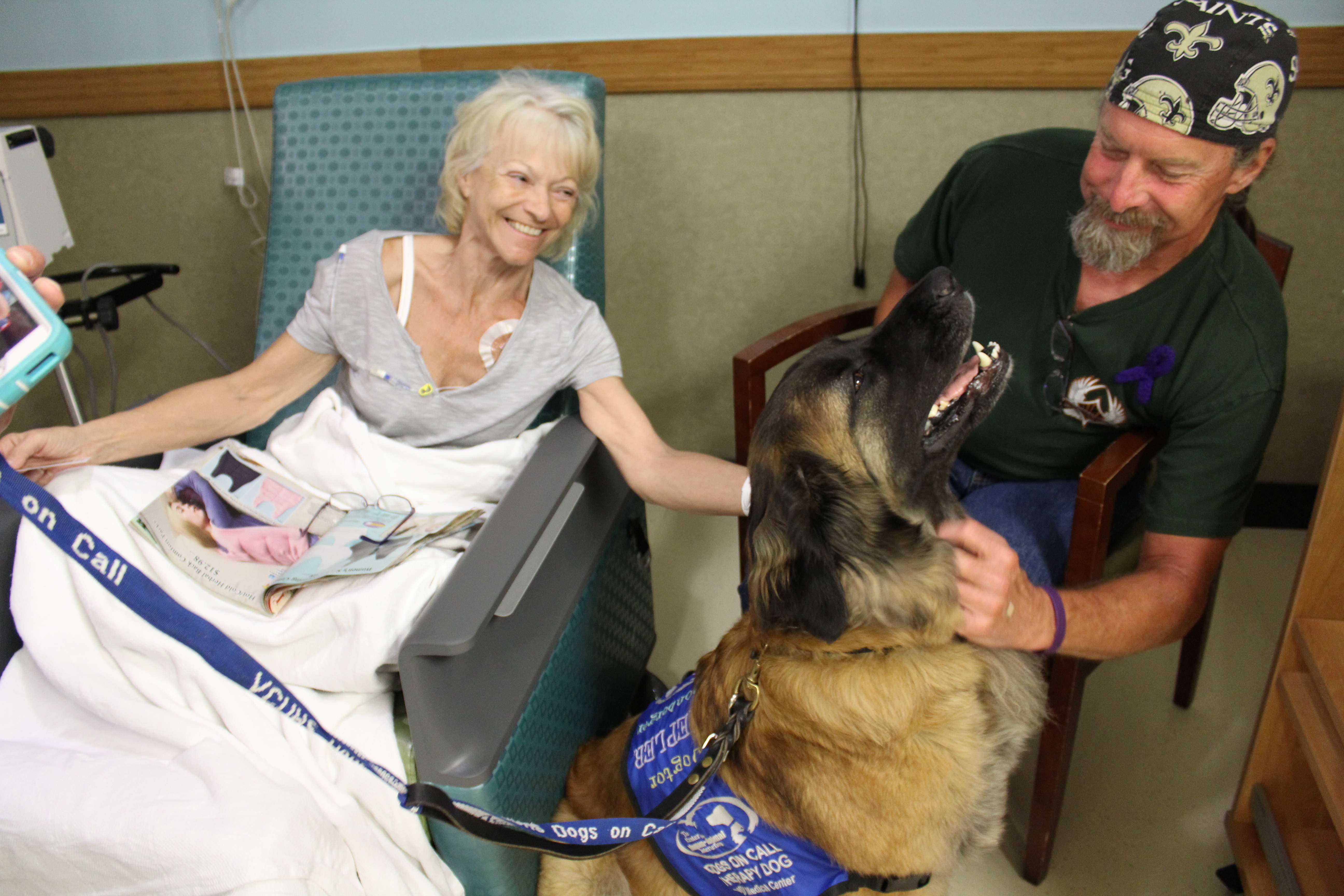 Dogs On Call therapy dog Kepler, a leonberger who is wearing a blue Dogs On Call vest, receives neck scratches from a smiling friend in a chair. Kepler is leaning back in contentment and a patient sitting beside Kepler is smiling and also petting Kepler's head.